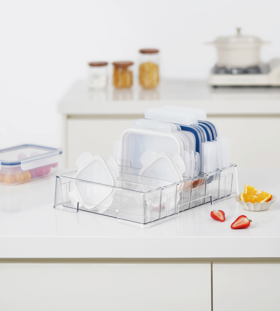 Auf einer Küchentheke steht der BNB x VCN Deckel Organizer KLAR - mit flexiblen Fächern, in dem Deckel und Behälter aus Kunststoff ordentlich aufbewahrt werden. In der Nähe befinden sich geschnittenes Obst, ein Behälter mit Deckel, und im Hintergrund Küchengläser und ein Topf.