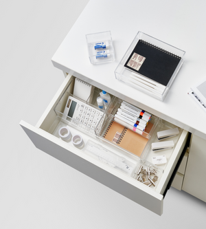 Eine offene weiße Schreibtischschublade ist mit dem SLIDE Schubladen Organizer KLAR - HOCH (BNB x VCN, verschiedene Größen) ordentlich geordnet und bietet Platz für die wichtigsten Büroutensilien wie Taschenrechner, Stifte, Textmarker, Klebeband, Büroklammern, Radiergummis und kleine Notizbücher; weitere Gegenstände befinden sich auf dem Schreibtisch darüber.