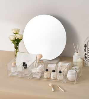 Ein Waschtisch mit rundem Spiegel, weißen Rosen in einer Vase, Hautpflege und Make-up im BNB x VCN SLIDE Schubladen Organizer KLAR - HOCH (verschiedene Größen), Pinsel, Wattepads, Diffusorstäbchen und Bücher auf der rechten Seite - alles auf einer beigen Fläche.