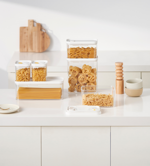 Auf der Küchentheke stehen stapelbare DATEMATE Vorratsdosen KLAR Weiss mit Garzeit-Timer von BNB x VCN mit Nudeln, gestapelte Schüsseln, hölzerne Salz- und Pfeffermühlen, ein Teller mit Gabel und Schneidebretter, die an der Wand lehnen.