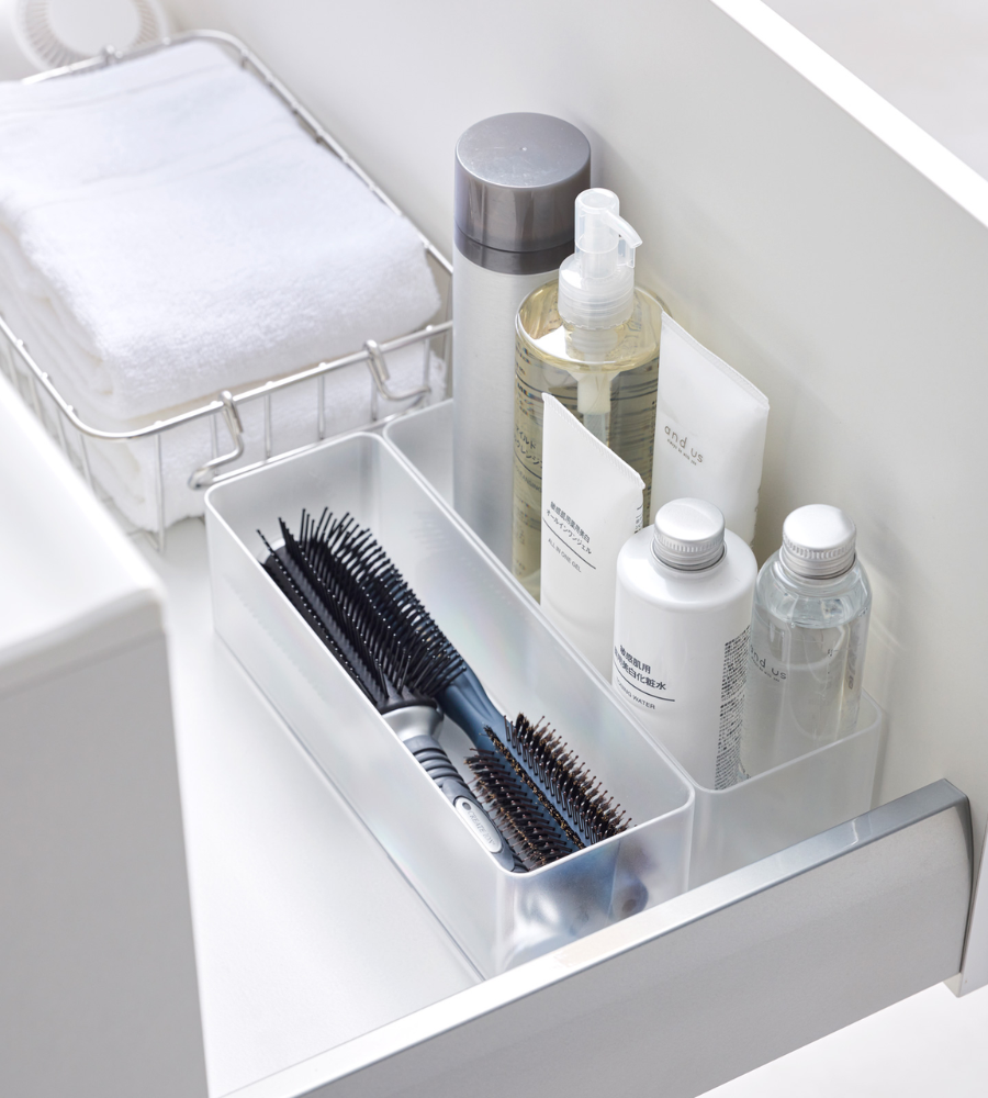 Eine geordnete Badezimmerschublade mit dem YAMAZAKI TOWER Schubladen Organizer Weiss L DEEP - Set 2 Stk. hält Haarbürsten, Flaschen, einen Metallkorb mit weißen Handtüchern und andere Pflegeartikel in durchsichtigen Behältern sauber.