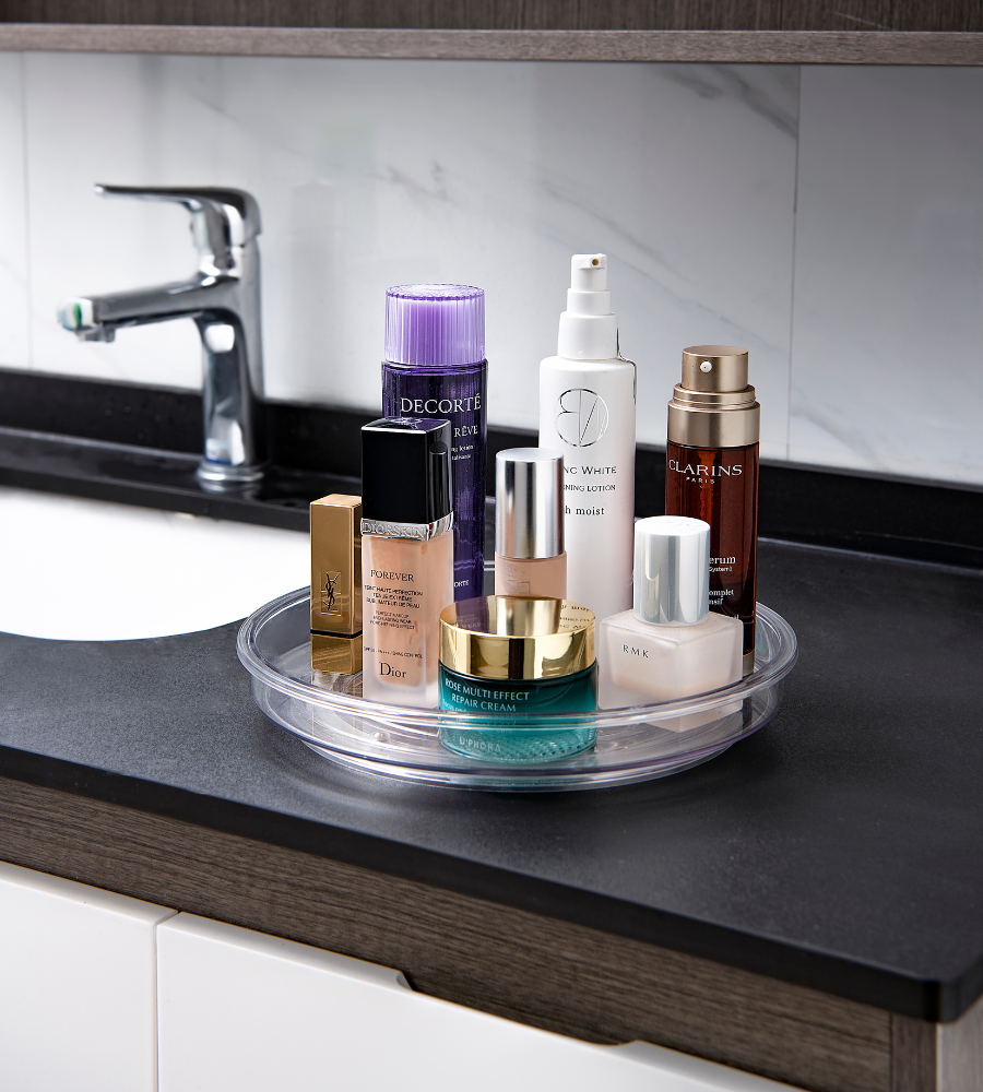 Die Drehteller Lazy Susan KLAR (23,5 x 3,7 cm) von BNB x VCN steht auf einem schwarzen Badezimmertisch und ordnet Hautpflege- und Kosmetikflaschen in der Nähe eines Waschbeckens mit silbernem Wasserhahn und Marmoraufkantung.