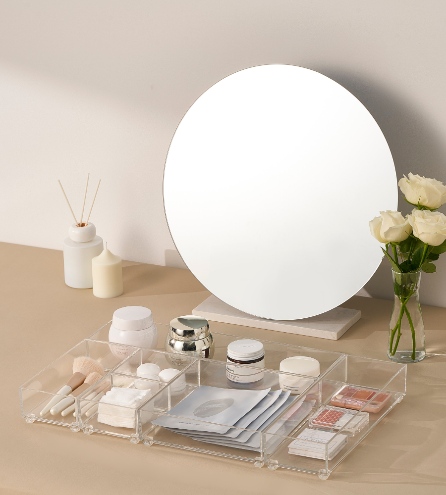 Ein runder Spiegel steht auf einem beigen Waschtisch mit weißen Rosen und Kerzen. Make-up-Pinsel, Cremes, Pflegeprodukte und Maskenblätter sind in einem Organizer und dem BNB x VCN Schubladen Organizer-Set SLIDE (5-teilig) geordnet.