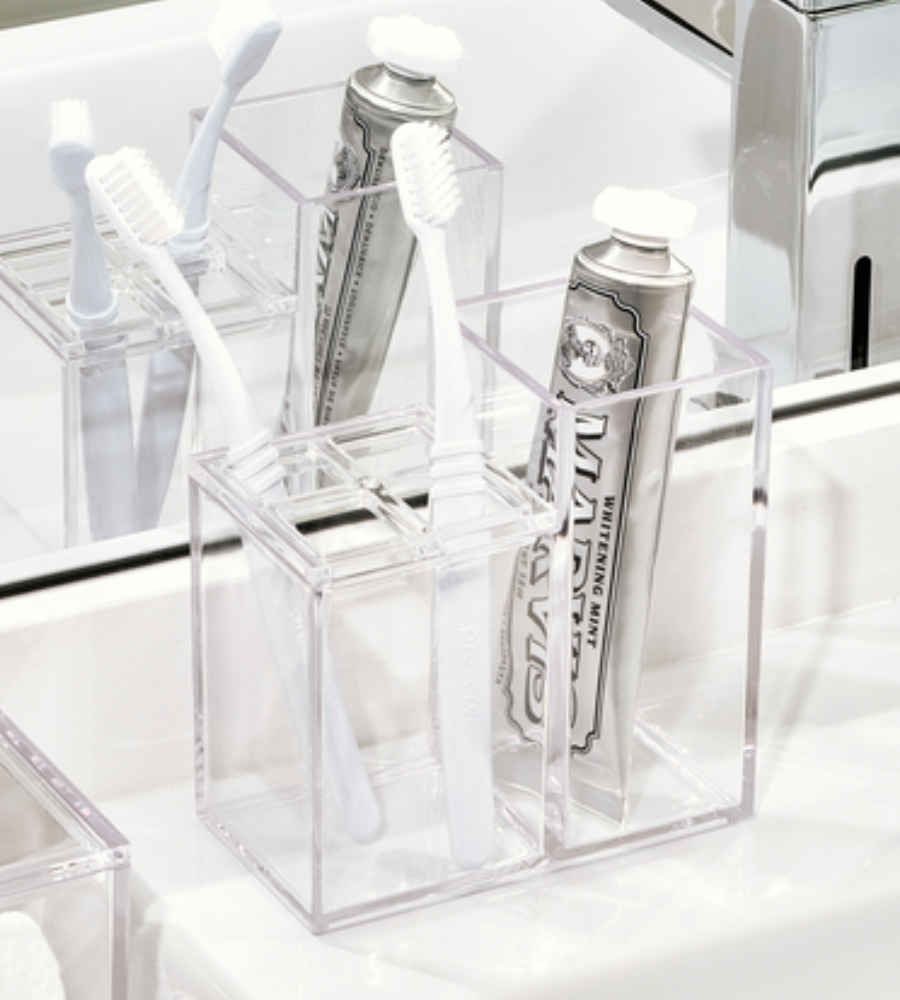Der iDesign CLARITY - Kosmetik Organizer KLAR II sitzt auf einem Badezimmer-Waschbecken und hält als Zahnbürstenhalter zwei weiße Zahnbürsten und eine silberne Zahnpastatube, deren Reflexion im dahinter liegenden Spiegel sichtbar ist.